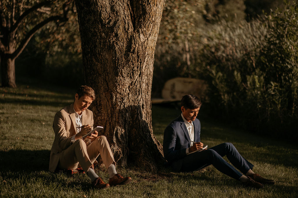LGBTQ+ couple during their elopement writing in notebooks while sitting against a tree.