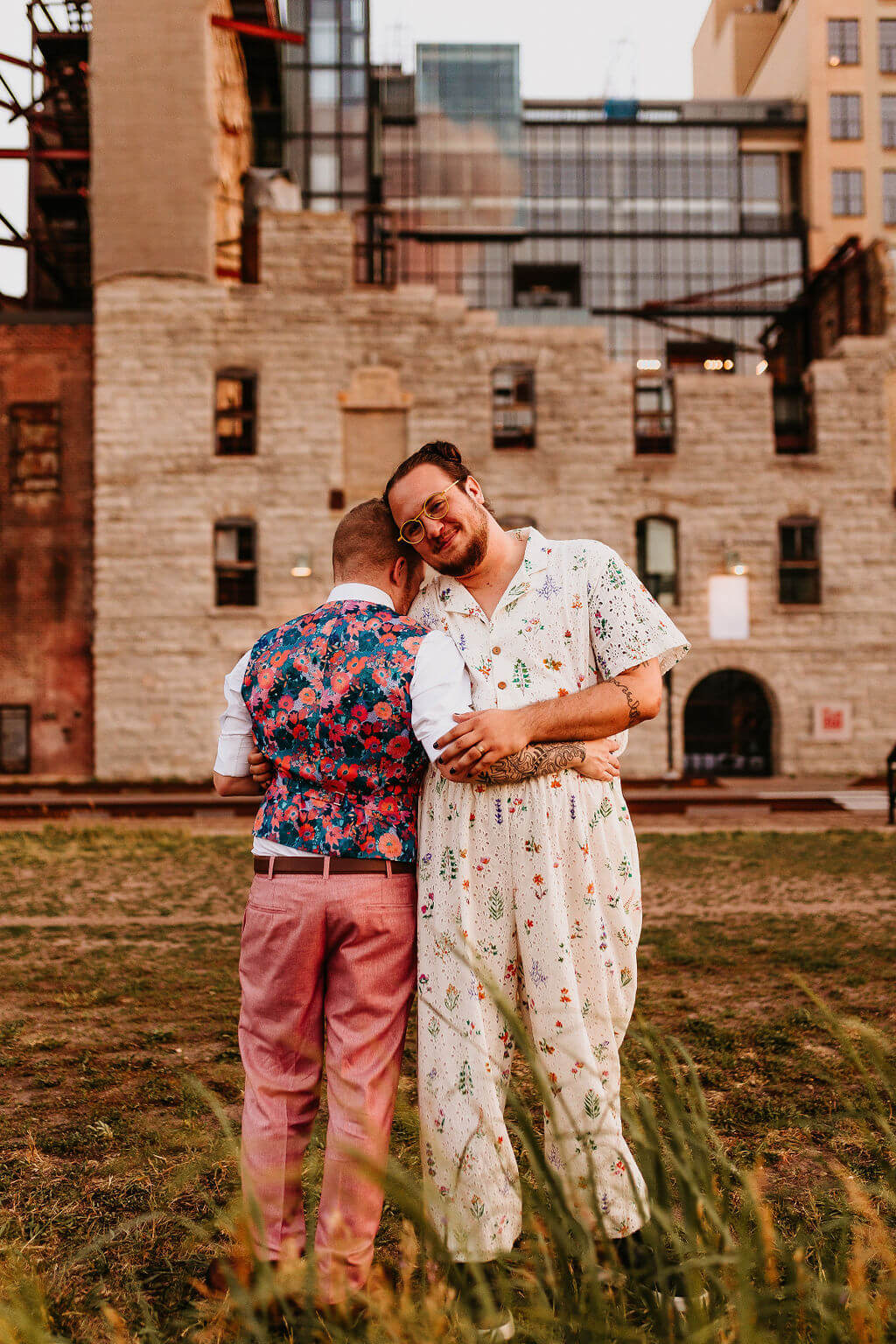 LGBTQ+ couple posing on their wedding day. One is in a jumpsuit and the other is in a suit with a flowery vest.