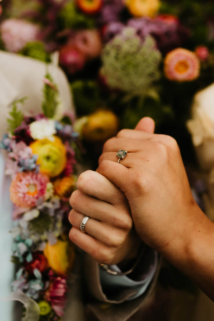 Close up of a queer couple holding hands with their rings in view and flowers in the background.